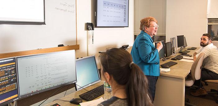 Professor Marian Extejt teaching a class standing at the front of the classroom flanked by two tables each with a student sitting at a computer